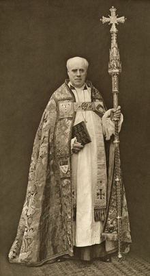 bald, clean-shaven white man in ecclesiastical robes, carrying as staff surmounted by a cross