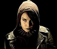 A young woman in a hooded black leather jacket and spiked choker, with heavy eye-makeup and black lipstick on, and piercings in her eyebrow and nose, looks coldly into the camera.