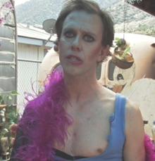 Sean Whalen in dim lighting, with heavy make-up around his eyes. He wears a light blue tank top with a pink scarf over it. His left nipple is exposed.