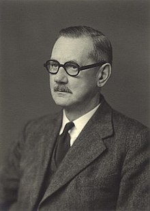 Allen, a middle-aged man with a moustache wearing round spectacles, sits in three-quarter profile.