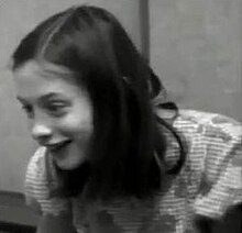 A black and white frame taken from a video of Genie, who is enthusiastically smiling, taken from several feet away. It shows her from the chest up, and Genie is facing slightly to the right of the camera.