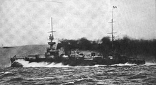 A large black ship, partially obscured by thick smoke from its funnels, creates a large bow wave as it plows through the water at high speed.