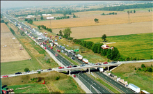 "Ontario Highway 401 viewed from high above travels into the distance from the bottom-right to the top-left. The Manning Road overpass crosses the highway near the bottom of the image. The surroundings are entirely agricultural. On the highway, several dozen vehicles are piled into each other. The middle of the large pileup is smoking."