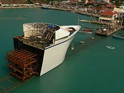A full-size model of a cruise ship's bow is seen from behind, supported by large frame. The model sits on top of a rail, which is underwater and leads underneath a marina pier. Boats are located in the water near the marina, and small buildings are in the background.