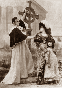 Tall white man, clean shaven, in rich ecclesiastical robes, and a young woman in period costumes who is protectively clutching a young child; they are at a graveside