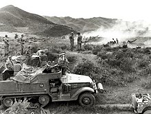 A half track vehicle sits in the foreground in a grassy open area, with a large-calibre machine gun mounted on top. Soldiers using binoculars are sitting on the vehicle, while in the background smoke obscures a number of artillery guns which are firing