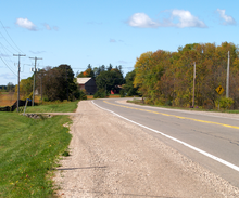 A two lane highway with gravel shoulders passes through farmland, with a barn in the distance