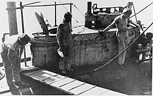 The conning tower of a submarine while docked. Four men stand around the conning tower and a gangway plank has been laid down next to it. The open hatch of the submarine and various ropes mooring the boat can also be seen.