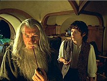 screenshot of pipe-smoking bearded Wizard and child-sized Hobbit in a miniature house with rounded ceilings