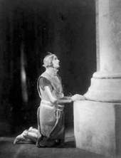 stage scene depicting a white woman wearing medieval armour, kneeling in a church