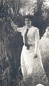 young woman in full-length frock and top coat leaning on a tree