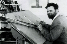 A bearded man in his mid-30s, dressed in a shirt and woolen pullover, is sitting at a drawing board. His pencil is poised in his right hand, and he is smiling off to the left of the camera.