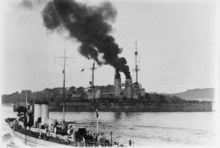 A two-funneled ship steams through a harbor with smoke billowing