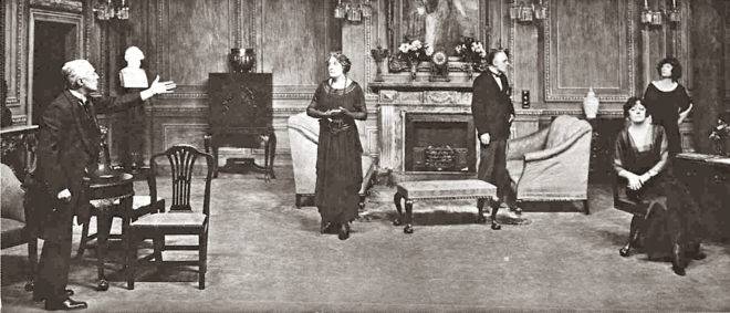 stage indoor scene: three men and two women in discussion, with some gesticulation