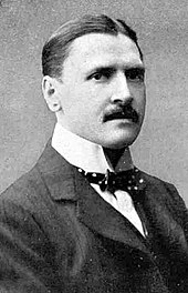 white man in early middle age; he has a full head of neat dark hair and a small moustache