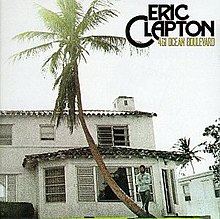 A house is shown along with a palm tree, on the right Eric Clapton holds his hands smiling, above him is the title and logo along with some trees