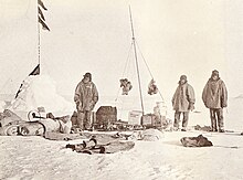 On the left is a snow cairn with flags. Three men are nearby, and assorted equipment is strewn on the snow.