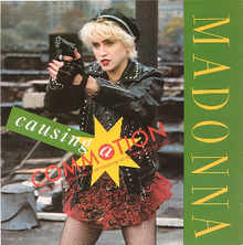 Madonna with short cropped blond hair is pointing towards somebody while holding a gun. She is wearing a red skirt, a black jacket and gloves.