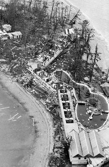 Black and white aerial photograph of a small island with unroofed buildings, trees stripped of their leaves and branches, and debris scattered over the ground and beach.