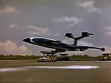 A gleaming, blue, futuristic airliner rests on top of three orange support vehicles (one underneath the fuselage and two underneath the wings) as it speeds out of control along a runway against a grassy backdrop, the sky bright with clouds.