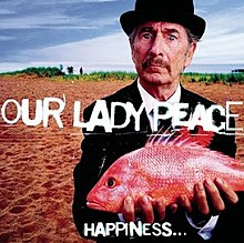 An old man standing on a sandy beach wearing a black suit and bowler derby stands holding an orange-pink fish in both hands. He stares at the audience with a puzzled look on his face.