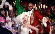 Image of several people at a party. On the left, there are people wearing colorful make-up and outfits as a woman and a man stand out on the middle; the woman wears a white dress, a hat and an eye-patch, while the man wears a red suit and glasses. Other people appear on their back as candles are also featured.