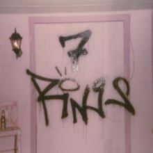 Graffiti on a pink wall, placed on the center of a dark pink door jamb, reading "7 RiNGS" with a dot over the letter I stylized as a ring. On the left side, there is a black lamp fixture and a pink chair with a bottle.