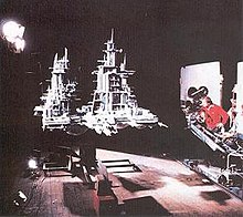 In a film studio, a director operates a crane-mounted camera aimed at a large gray model spacecraft, raised several feet in the air by a support structure underneath. The model is lit from one side by bright studio lights and has tall spires with many pieces protruding from them.