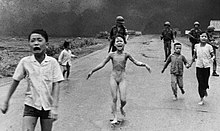 Scared children flee on a road, with soldiers behind them and a smoky sky; in the center is a nude girl, screaming and lifting her arms while running