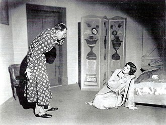 young man in a dressing gown looking down with a horrified expression at a middle-aged woman slumped to her knees on a bedroom floor looking back at him