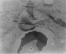 Heurtley, in a shirt, trousers and wide-brimmed hat, sitting in an excavation trench next to a large ancient pot.