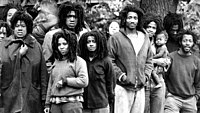 A black and white photo of group of around a dozen people standing outside. Everyone in the photo is black and has the same style of dreadlocks. The ages of the group vary, from a baby being held in someone's arms to a man who could be in his twenties or thirties.