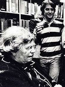 Alan H. Goodman standing beside Margaret Mead, famous Anthropologist. The image is in black and white. A young Alan, a white man in a striped shirt with hair to his chin, is standing behind Margaret Mead, an older woman with short hair. Both are in front of bookshelves.