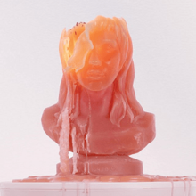 A red-orange candle representing Kesha's head is positioned on top of a surface. The candle wax is melted, disfiguring the top of the "head".