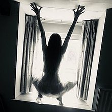 Silhouette of the back of a woman crouched before a window, with hands outstretched above her, pressed against a low ceiling