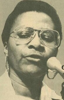 A Black woman with short dark hair, wearing aviator glasses and hoop earrings, speaking into a microphone