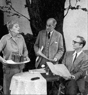 elderly white woman standing, holding a tray, middle aged white man standing next to her, and middle aged white man, bespectacled, sitting at a table, holding a newspaper