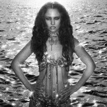 A black-and-white photograph of Jess Glynne standing in front of water with her hands on her hips wearing a sheer dress