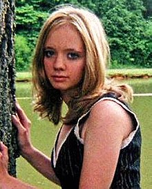 A Caucasian woman with blonde shoulder-length hair wearing a blue tank top with white trim looks to her left directly at the camera with her hands placed on a tree at the left of the image. Behind her is a body of water and some woods.