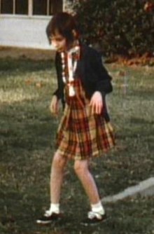 A slightly blurry color picture of Genie, facing slightly right of the camera, walking by herself outside in the hospital recreation yard. She is wearing a red and Orange plaid-patterned dress and thin blue sweater and looks extremely pale, emaciated, and expressionless. Her limbs are exposed and look extremely thin. Both of her knees are very bent, and her arms are bent forward with both hands hanging down as she holds them out in front of her.