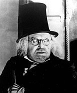 A close-up of a man with a black coat, round-framed glasses, a tall black top hat, and dissheveled white hair. The man is looking toward the right out of the picture.